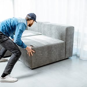 Courier or handyman in blue shirt and cap mounting a new couch in the apartment