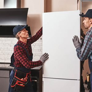 Moving again. men mechanics moving refrigerator to another place. They assist each other. Side view